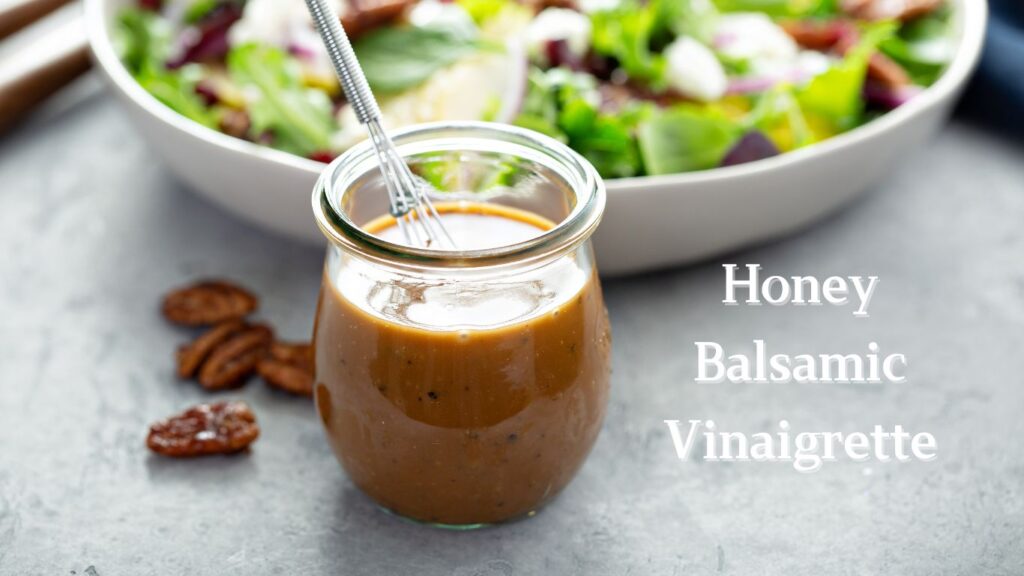 A jar of honey balsamic vinaigrette with a spoon inside, next to a bowl of mixed greens salad.”