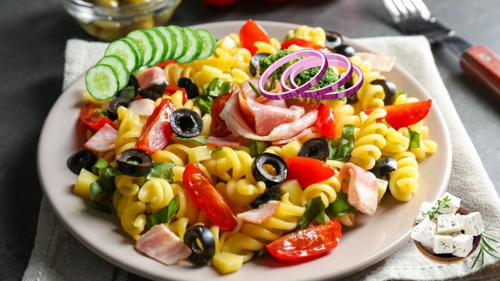 Plate of vibrant summer pasta salad with cucumbers, red onions, black olives, cherry tomatoes, and ham slices.