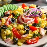 Plate of vibrant summer pasta salad with cucumbers, red onions, black olives, cherry tomatoes, and ham slices.