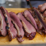 Slices of smoked brisket being cut on a wooden board by a chef's hand with a knife.