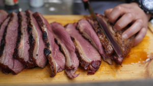 Slices of smoked brisket being cut on a wooden board by a chef's hand with a knife.