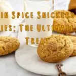 A display of pumpkin spice snickerdoodle cookies with a glass of milk, cinnamon sticks, and pumpkins in the background.