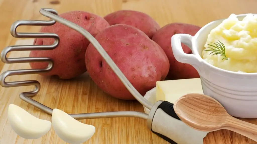 A close-up image of red potatoes, a garlic clove, a pat of butter, a wooden spoon, and a bowl of mashed potatoes garnished with dill on a wooden surface.