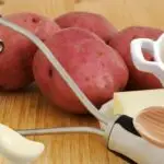 A close-up image of red potatoes, a garlic clove, a pat of butter, a wooden spoon, and a bowl of mashed potatoes garnished with dill on a wooden surface.