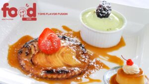 A plate featuring a caramel-drizzled apple pie topped with a strawberry, a small ramekin of green pudding garnished with a blackberry, and a flan with whipped cream and a cherry.