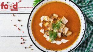 A bowl of creamy pumpkin soup topped with grilled chicken pieces, fresh dill, and a drizzle of cream, served in a white bowl with black polka dots.