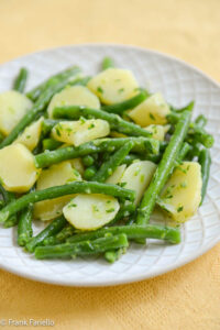 A colorful and decorative image featuring a green bean and potato salad. The image includes various elements such as green beans, peas, and red potatoes, along with vibrant hearts, stars, and a butterfly. The text "GREEN BEAN AND POTATO SALAD" is prominently displayed in green font. There is also a "read more" button with a smiling face and the text "Tom Tuesday Dinner" at the bottom.
