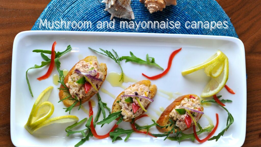 A white rectangular plate with mushroom and mayonnaise canapés garnished with red and green strips of bell pepper, purple onion slices, and lemon wedges.