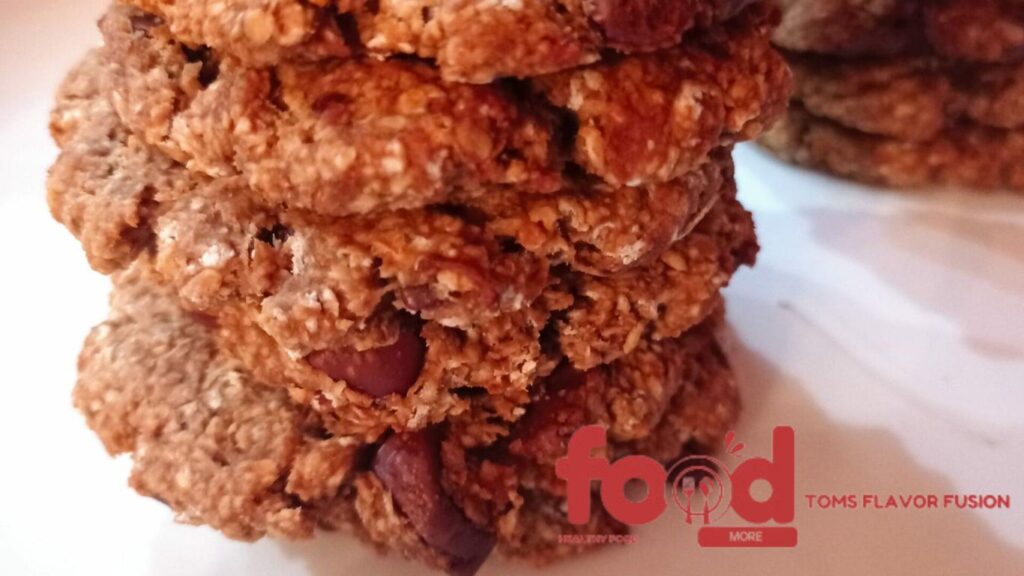 Close-up of a stack of oatmeal chocolate chip cookies with a logo in the bottom right corner that reads "food" with a spoon and fork icon, and "TOMS FLAVOR FUSION" in smaller text.