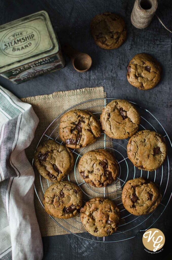 A batch of chocolate chip cookies cooling on a wire rack, surrounded by rustic kitchen items including a vintage tin, a wooden scoop, and a spool of twine.