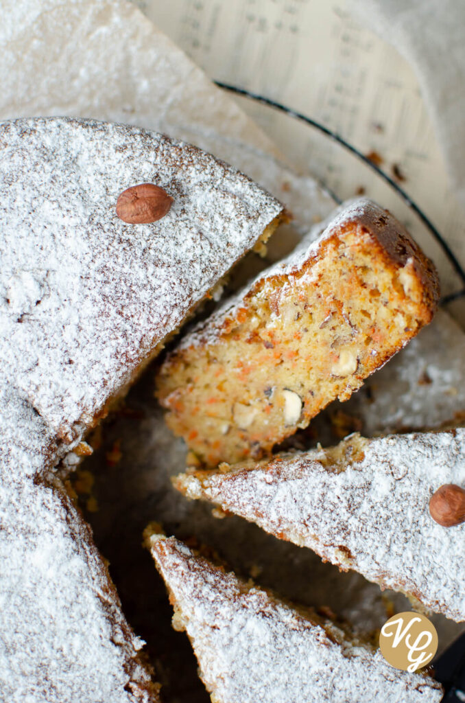 A close-up of a sliced carrot cake dusted with powdered sugar and garnished with whole hazelnuts.