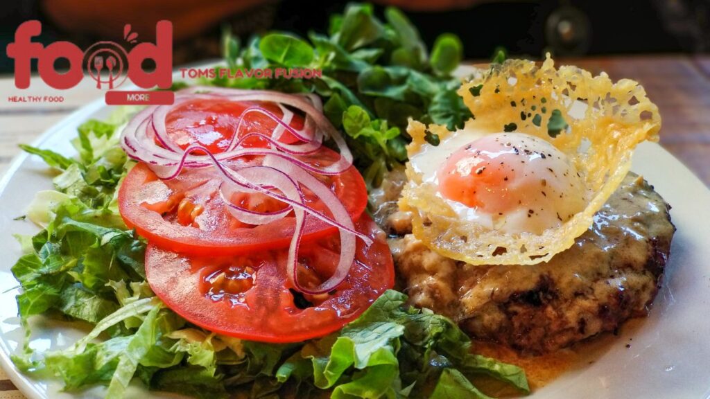 Homemade hamburger steak topped with a poached egg and crispy cheese, served with fresh tomatoes, red onions, and leafy greens.