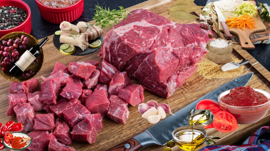 A wooden cutting board with various ingredients including a large piece of raw beef, diced beef, garlic cloves, a bowl of tomato paste, olive oil, sliced tomatoes, a knife, and various spices and herbs.