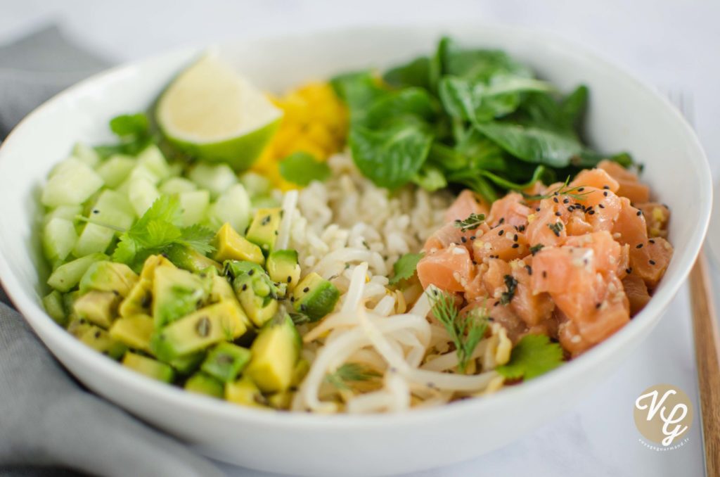 A bowl of fresh ingredients including diced cucumber, avocado, lime wedge, leafy greens, diced salmon, rice, and noodles, garnished with herbs and black sesame seeds.