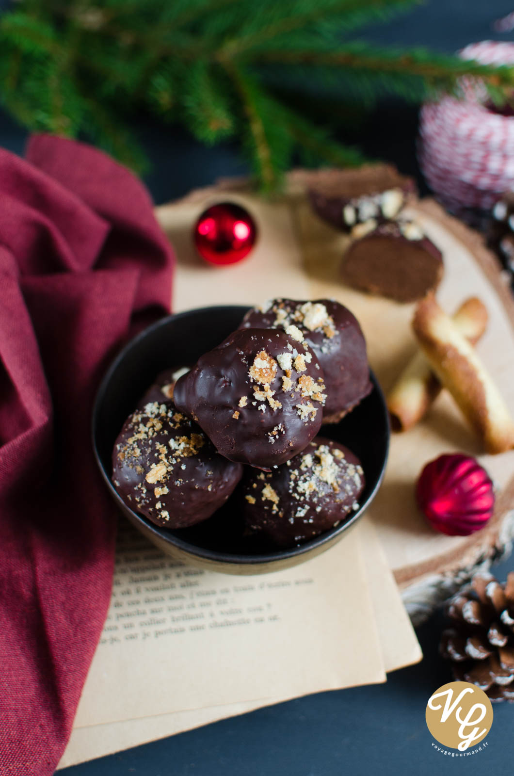 A bowl of chocolate-covered pralines topped with crushed nuts, surrounded by festive decorations including red ornaments, pinecones, and a red cloth.