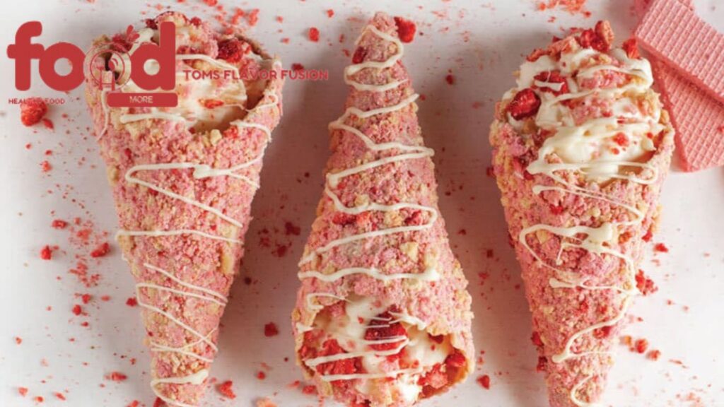 Three strawberry shortcake cones with white chocolate drizzle on a white surface, surrounded by pink crumbs.