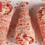 Three strawberry shortcake cones with white chocolate drizzle on a white surface, surrounded by pink crumbs.