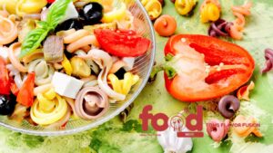 A colorful bowl of gluten-free Italian pasta salad featuring vibrant tri-color pasta, fresh basil, olives, tomatoes, bell peppers, and cheese cubes, placed next to a bundle of fresh basil leaves on a textured gray surface.