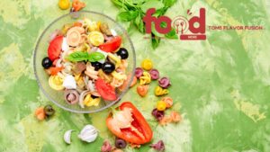  A colorful bowl of gluten-free Italian pasta salad featuring vibrant tri-color pasta, fresh basil, olives, tomatoes, bell peppers, and cheese cubes, placed next to a bundle of fresh basil leaves on a textured gray surface.