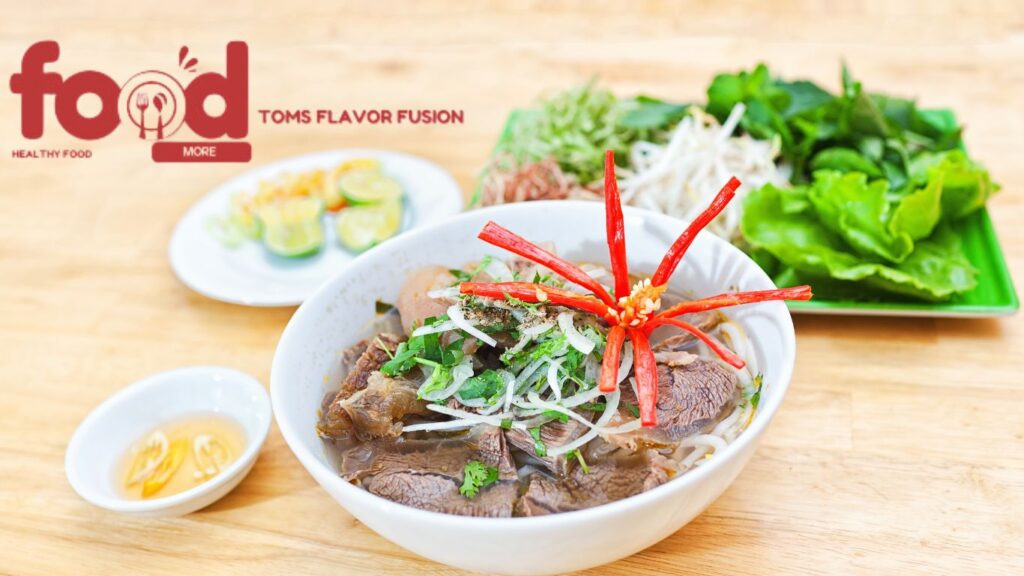 A bowl of Vietnamese beef noodle salad (Bun Bo Xao) garnished with fresh herbs and sliced red chili peppers, accompanied by plates of fresh vegetables and lime wedges.