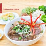 A bowl of Vietnamese beef noodle salad (Bun Bo Xao) garnished with fresh herbs and sliced red chili peppers, accompanied by plates of fresh vegetables and lime wedges.