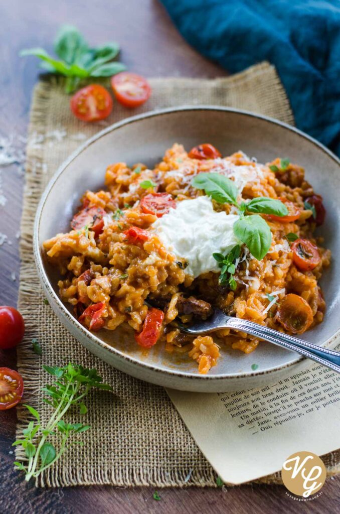 A beautifully plated dish of creamy risotto topped with candied tomatoes, crispy smoked bacon, and creamy burrata, garnished with fresh basil leaves.