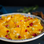 A close-up of a plate of sliced oranges garnished with pomegranate seeds and chopped pistachios.