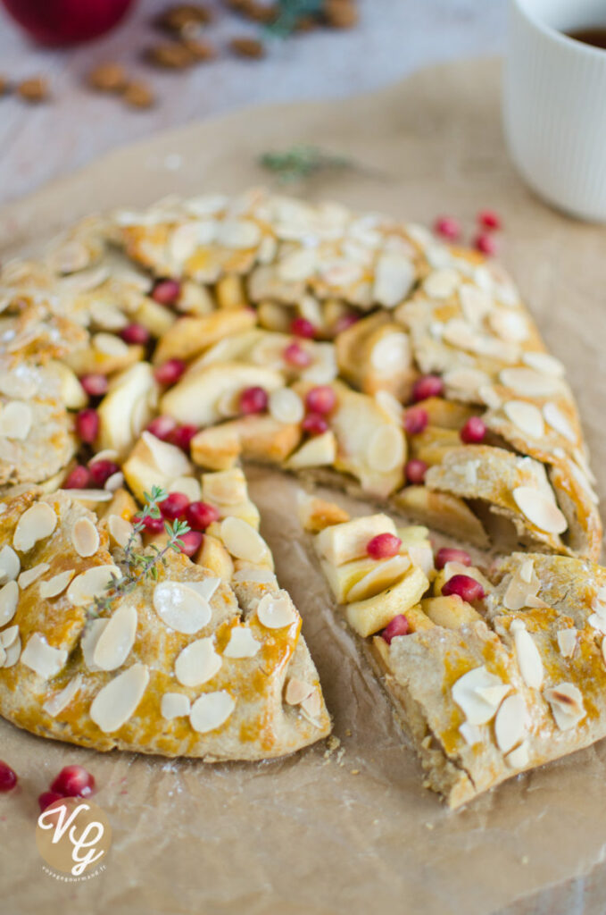 A close-up of a rustic galette topped with almond slices, apple slices, and pomegranate seeds, with a slice cut out. The galette is placed on a piece of parchment paper, and a white cup is partially visible in the background.