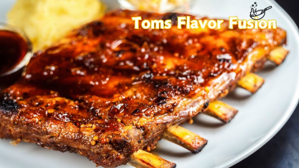 A plate of glazed barbecue ribs with a side of mashed potatoes and a small bowl of sauce. The text "Toms Flavor Fusion" is written in yellow at the top right corner of the image with a small illustration of a frying pan.