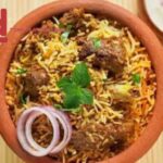 A clay pot filled with biryani garnished with mint leaves and onion rings, accompanied by bowls of raita and curry.