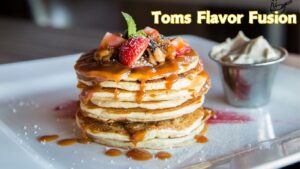 A stack of pancakes topped with strawberries, nuts, and caramel sauce, with a side of whipped cream in a metal cup. The text "Toms Flavor Fusion" is displayed in the top right corner.