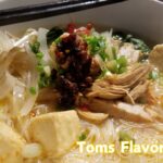 A close-up of a flavorful bowl of creamy chicken noodle soup with shredded chicken, tofu, green onions, bean sprouts, and chili oil, served with chopsticks and a spoon.