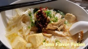 A close-up of a flavorful bowl of creamy chicken noodle soup with shredded chicken, tofu, green onions, bean sprouts, and chili oil, served with chopsticks and a spoon.