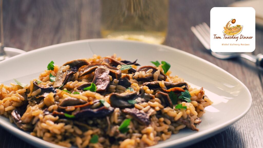 A close-up of a plate of mushroom risotto garnished with fresh herbs, with a glass of white wine in the background and a logo for "Tom Tuesday Dinner" in the top right corner.