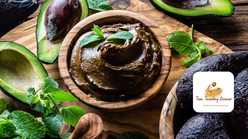 A wooden bowl filled with a dark brown creamy dip, garnished with fresh mint leaves, surrounded by halved avocados and fresh mint sprigs on a wooden surface. The logo of "Tom Tuesday Dinner - Global Culinary Recipes" is visible in the bottom right corner.