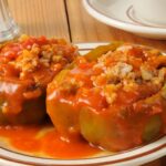 Two stuffed green bell peppers topped with tomato sauce on a white plate with a brown rim, placed on a wooden table. Decorative elements including flowers, geometric shapes, and a cartoonish face with large eyes are added around the image.