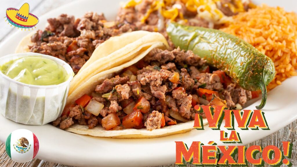 A plate of Mexican food featuring two tacos filled with seasoned ground beef, diced onions, and tomatoes, accompanied by a grilled green chili pepper, a serving of rice, and a small cup of guacamole. The text "¡Viva la México!" is prominently displayed in the bottom right corner, with a small Mexican flag and a sombrero graphic in the top left corner.