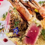 A gourmet dish featuring seared tuna tataki coated in sesame seeds, paired with plump grilled shrimp, served on a bed of fluffy couscous and garnished with fresh herbs and edible flowers.