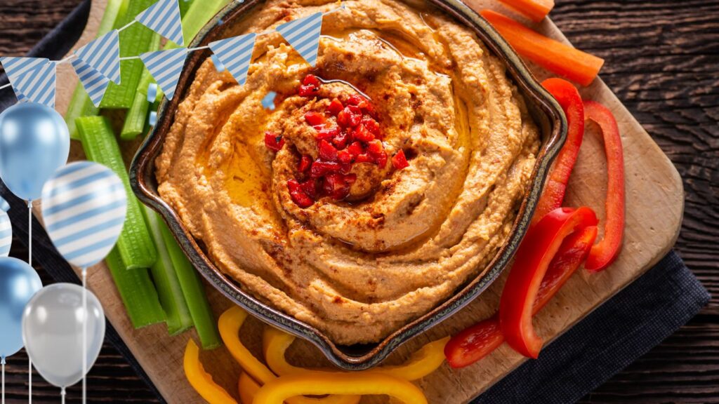 A bowl of hummus garnished with chopped red peppers, surrounded by celery sticks and sliced bell peppers on a wooden cutting board, with blue and white party decorations including balloons and bunting.