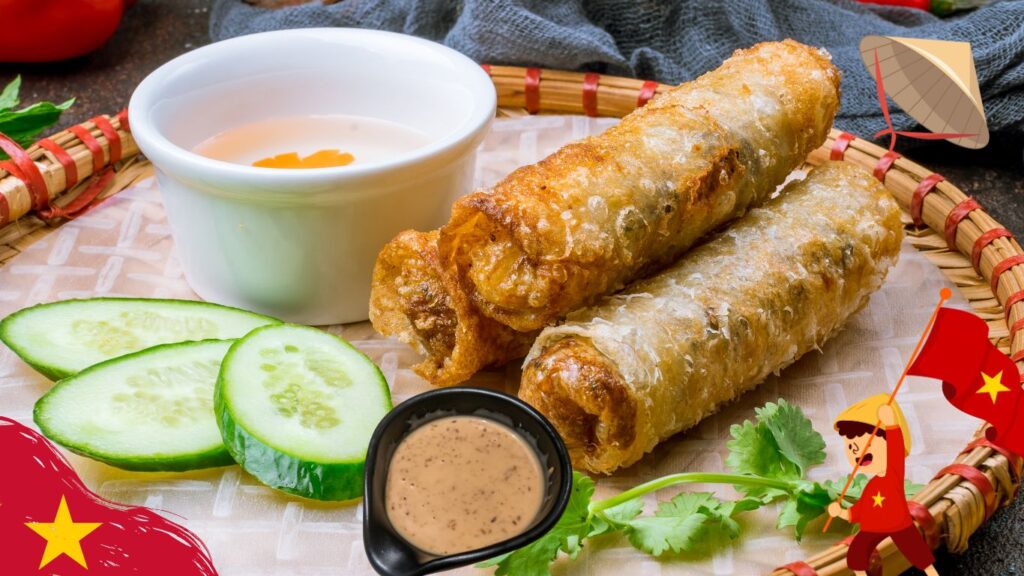 A close-up of three crispy Vietnamese spring rolls served on a woven bamboo tray with a small bowl of dipping sauce, cucumber slices, and a small dish of creamy sauce. The tray is decorated with a Vietnamese flag and a cartoon character holding a flag.