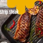 Two grilled strip steaks on a grill pan with garlic, red chili peppers, thyme, and seasoning, presented on a rustic wooden table.