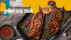 Two grilled strip steaks on a grill pan with garlic, red chili peppers, thyme, and seasoning, presented on a rustic wooden table.