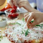 Hands adding fresh arugula to homemade pizza with melted cheese and tomato sauce on parchment paper