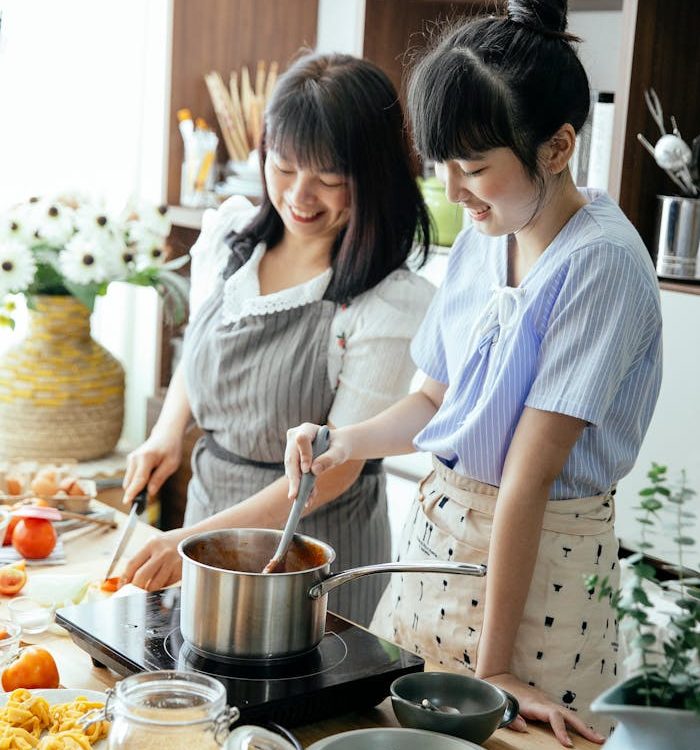 Cheerful Asian woman with young daughter smiling while cutting veggies and stirring sauce during lunch preparation together in kitchen